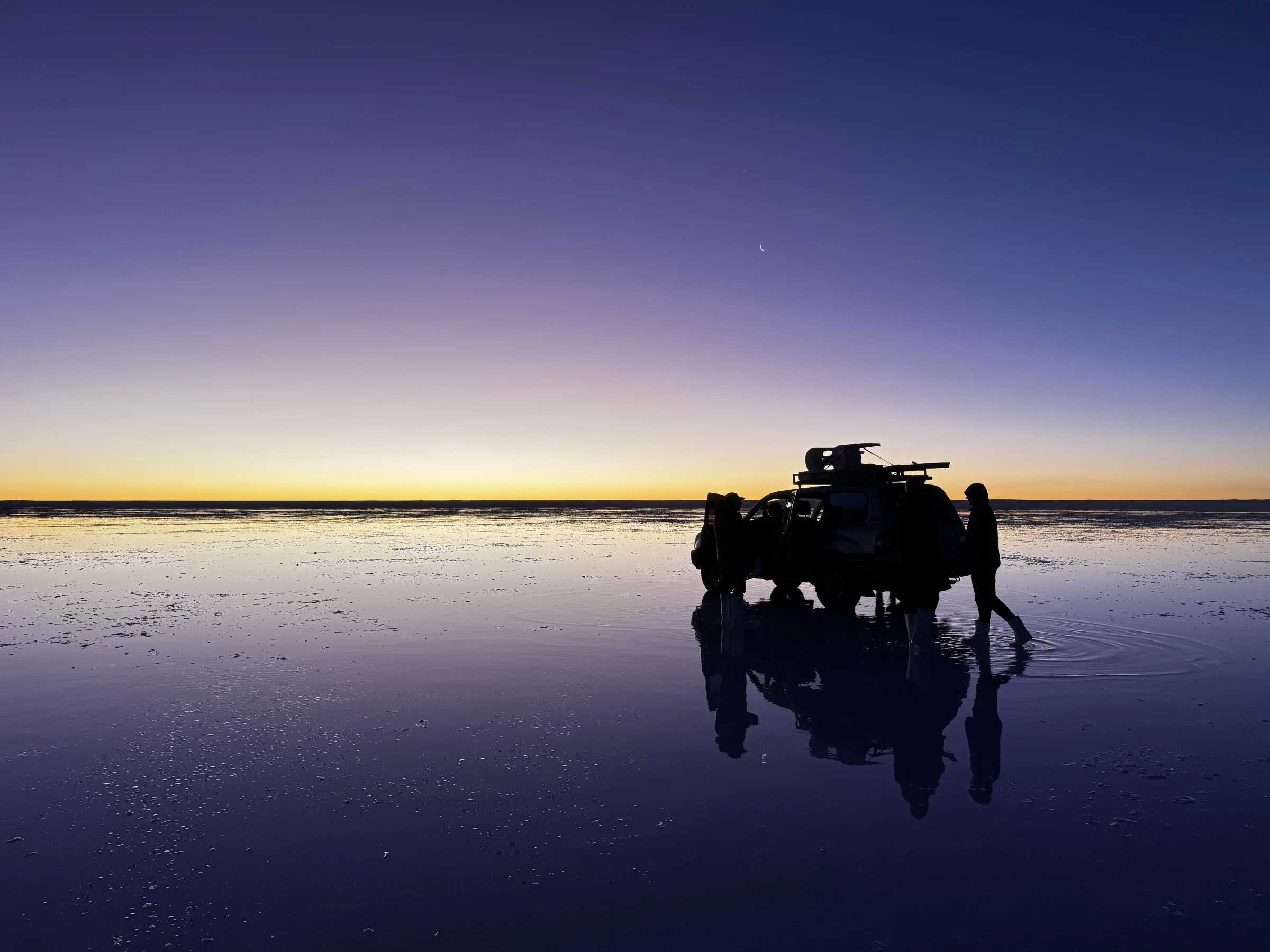 Travellers beside a four-wheel-drive vehicle reflected in Salar de Uyuni at twilight